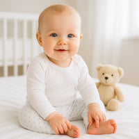 Baby sitzt auf hellem Bett und trägt weiße Baby Corner Hose mit blauem Krönchen-Print – atmungsaktive Baumwolle, Größe 56–62; bequem für Alltag und Spielzeit, süßer Look mit Teddy im Hintergrund.
