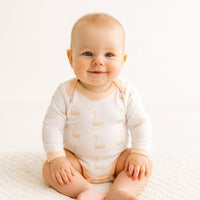 Weißer Fresk Langarm-Body mit Schwanenmotiv am Baby, sitzend im hellen Babyzimmer – aus 100% Bio-Baumwolle, bequem und alltagstauglich; sanfte Farben, unisex; schönes Geschenk zur Geburt und vielseitig kombinierbar.