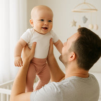 Mellow-rose Babyhose von Fresk am Baby im Kinderzimmer, Papa hebt das Kind lachend hoch – weiche Bio-Baumwolle, komfortabler Bund, vielseitig kombinierbar für Alltag und Erstausstattung.