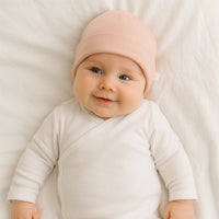 Baby trägt Fresk Babymütze in mellow rose und liegt auf weißer Decke – kuschelige Bio-Baumwolle, hält warm und sitzt bequem; schöne Ergänzung zur Erstausstattung.