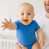 Baby im blauen Name it Kurzarm-Body streckt lachend die Hand aus und sitzt auf Papas Arm – atmungsaktive Baumwolle, Größe 56; pflegeleicht und alltagstauglich.