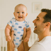 Papa hält lachendes Baby im weißen Name it Kurzarm-Body mit blauen Traktoren – atmungsaktive Baumwolle, Größe 56; bequem für Alltag und Spielzeit.