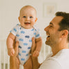 Papa hält lachendes Baby im weißen Name it Kurzarm-Body mit blauen Traktoren – atmungsaktive Baumwolle, Größe 56; bequem für Alltag und Spielzeit.