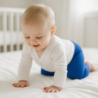 Baby krabbelt auf weißem Bett und trägt blaue Playshoes Babyleggings in Größe 50–56 – weiche Erstausstattung für Neugeborene.