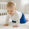 Baby krabbelt auf weißem Bett und trägt blaue Playshoes Babyleggings in Größe 50–56 – weiche Erstausstattung für Neugeborene.