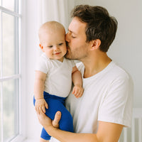 Vater hält sein Baby am Fenster; Baby trägt blaue Playshoes Babyleggings in Größe 50–56 – gemütliche Erstausstattung fürs Kinderzimmer.