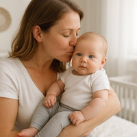 Mutter küsst ihr Baby im hellen Kinderzimmer; Baby trägt graue Playshoes Babyleggings in Größe 50–56 – weiche Erstausstattung.