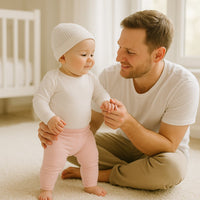 Baby sitzt im hellen Kinderzimmer und spielt, trägt rosa Babyleggings von Playshoes, Größe 50–56 – weiche Erstausstattung für Neugeborene.