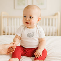 Baby sitzt auf dem Bett und trägt rote Playshoes Babyleggings in Größe 62–68 – weiche, bequeme Erstausstattung für Babys.