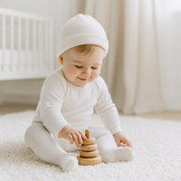 Baby sitzt im hellen Kinderzimmer und spielt, trägt weiße Playshoes Babystrumpfhose in Größe 50–56 – weiche Erstausstattung für Neugeborene.