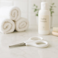 Weiße Playshoes Baby-Nagelschere mit abgerundeten Spitzen liegt auf dem Badezimmer-Waschtisch neben Handtüchern und Babylotion – sichere, hygienische Nagelpflege für Neugeborene und Babys.