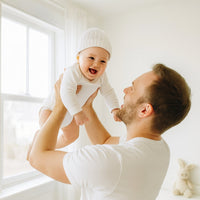 Papa hebt lachendes Baby im weißen Sanetta Langarm-Body – GOTS-zertifizierte Bio-Baumwolle, weich und pflegeleicht; Größe 56, ideal für Alltag und Erstausstattung.