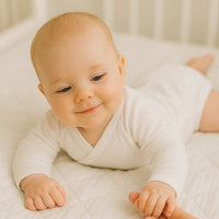 Baby in weißem Sanetta Wickelbody (langarm) liegt in Bauchlage auf hellem Bett und hält Mamas Finger – GOTS-zertifizierte Bio-Baumwolle, hautfreundlich und bequem, Größe 62.
