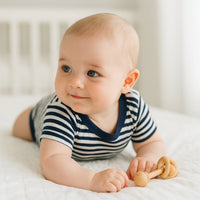 Baby in blau-grau gestreiftem Kurzarmbody von Schnizler in Bauchlage auf dem Bett; weiche Baumwolle und bequemer Sitz dank amerikanischem Ausschnitt – ideal für Alltag, Spielzeit und als Geschenk zur Geburt.
