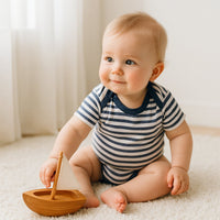 Baby trägt den kurzärmligen Schnizler Body in blau-weissem Ringelmuster und spielt im Kinderzimmer mit einem Holzboot – weiche Baumwolle, bequemer Schnitt mit amerikanischem Ausschnitt und Druckknöpfen; ideal für Neugeborene, hautfreundlich dank OEKO-TEX Klasse 1, Größe 50/56.