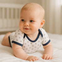 Baby trägt grau/blauen Schnizler Kurzarm-Body mit Sternen und liegt in Bauchlage auf hellem Bett – atmungsaktiver Baumwoll-Body, Größe 50–56; perfekt für warme Tage und den Alltag.
