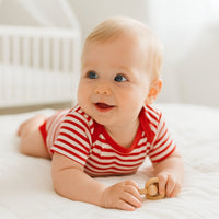 Baby trägt rot-weiß gestreiften Schnizler Kurzarm-Body und liegt auf hellem Bett – bequemer Baumwoll-Body Größe 50–56 für warme Tage und den Alltag.
