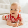 Baby trägt rot-weiß gestreiften Schnizler Kurzarm-Body und liegt auf hellem Bett – bequemer Baumwoll-Body Größe 50–56 für warme Tage und den Alltag.