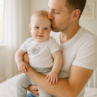 Graue Schnizler Nicki-Babyhose an lächelndem Baby im Arm des Papas; kuschelig, elastischer Bund und Bündchen für sicheren Sitz – hautfreundlich und OEKO-TEX zertifiziert, perfekt für jeden Tag.