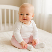Baby sitzt auf hellem Bett und trägt rosa gestreifte Schnizler Babyhose – bequeme Interlock-Baumwolle, Größe 62; gemütlich für Alltag und Spielzeit.