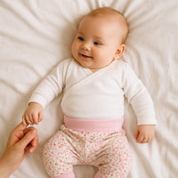 Rosa Schnizler Babyhose mit Blumenprint an Baby, liegend auf dem Bett – weiche Baumwolle, bequemer hoher Bund; ideal für Alltag, Krabbeln und als süßes Geschenk zur Geburt.