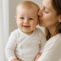 Baby im naturfarbenen Schnizler Wickelbody (langarm) im Arm der Mama, liebevolle Kuss-Szene – weiche Interlock-Baumwolle, Größe 56; gemütlich für Alltag, Schlaf und Wickelzeit.