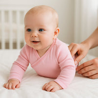 Lachendes Baby im rosa Schnizler Wickelbody mit langen Ärmeln, krabbelt/liegt auf dem Bett – kuschelige 100% Baumwolle (Interlock), Größe 56; bequemer Alltagsbody und schönes Geschenk zur Geburt.